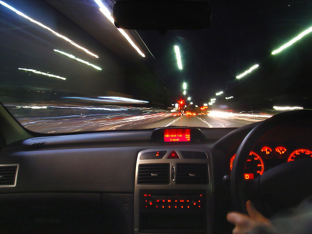 z pohledu auta je vidět rozmazaná cesta a rychlá světla, volant na pravé straně i budíky
